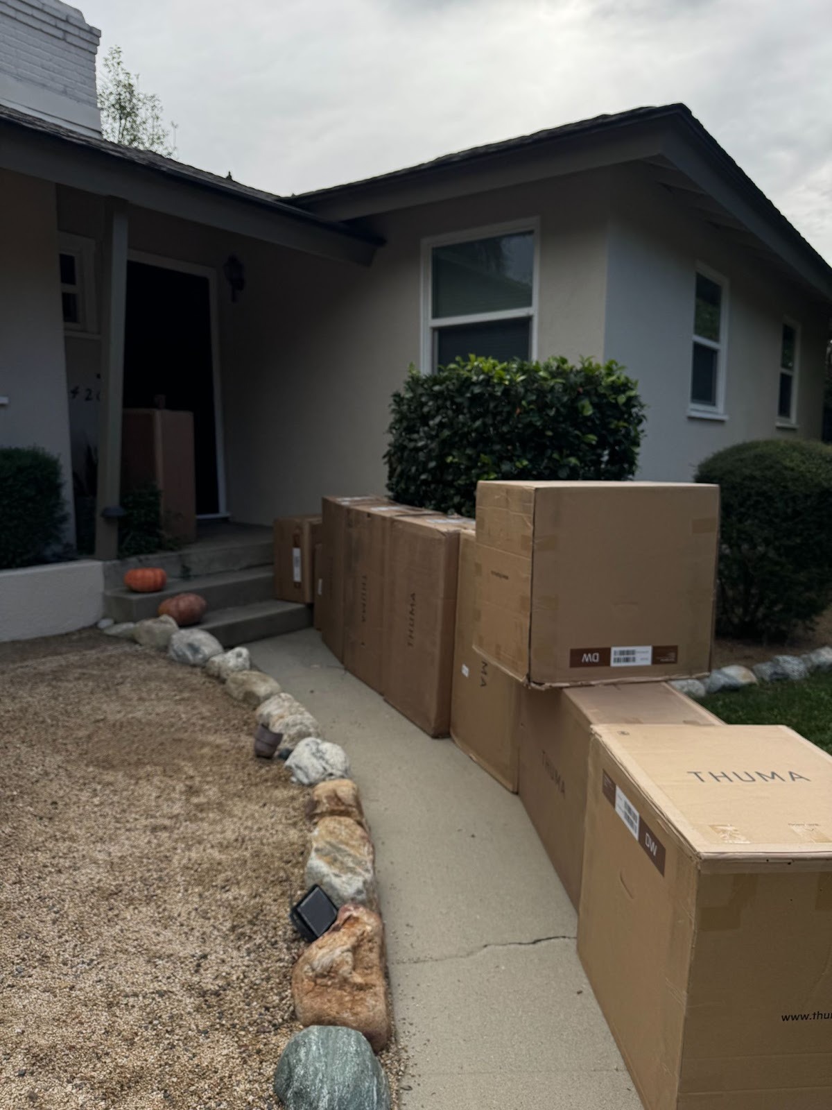 Several large cardboard boxes labeled "THUMA" are lined up along a walkway leading to the entrance of a house on a cloudy day.