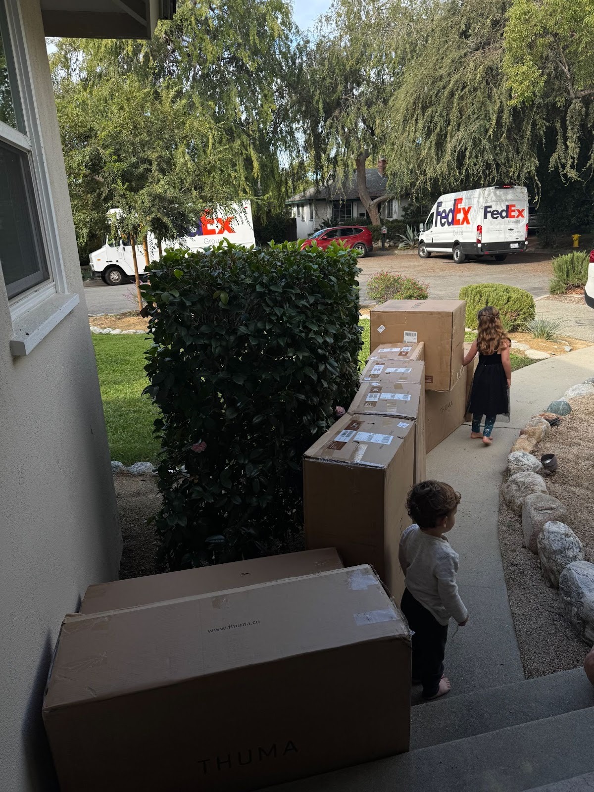 Two children stand near several large Thuma boxes on a front porch, with two FedEx trucks parked on the residential street in the background.