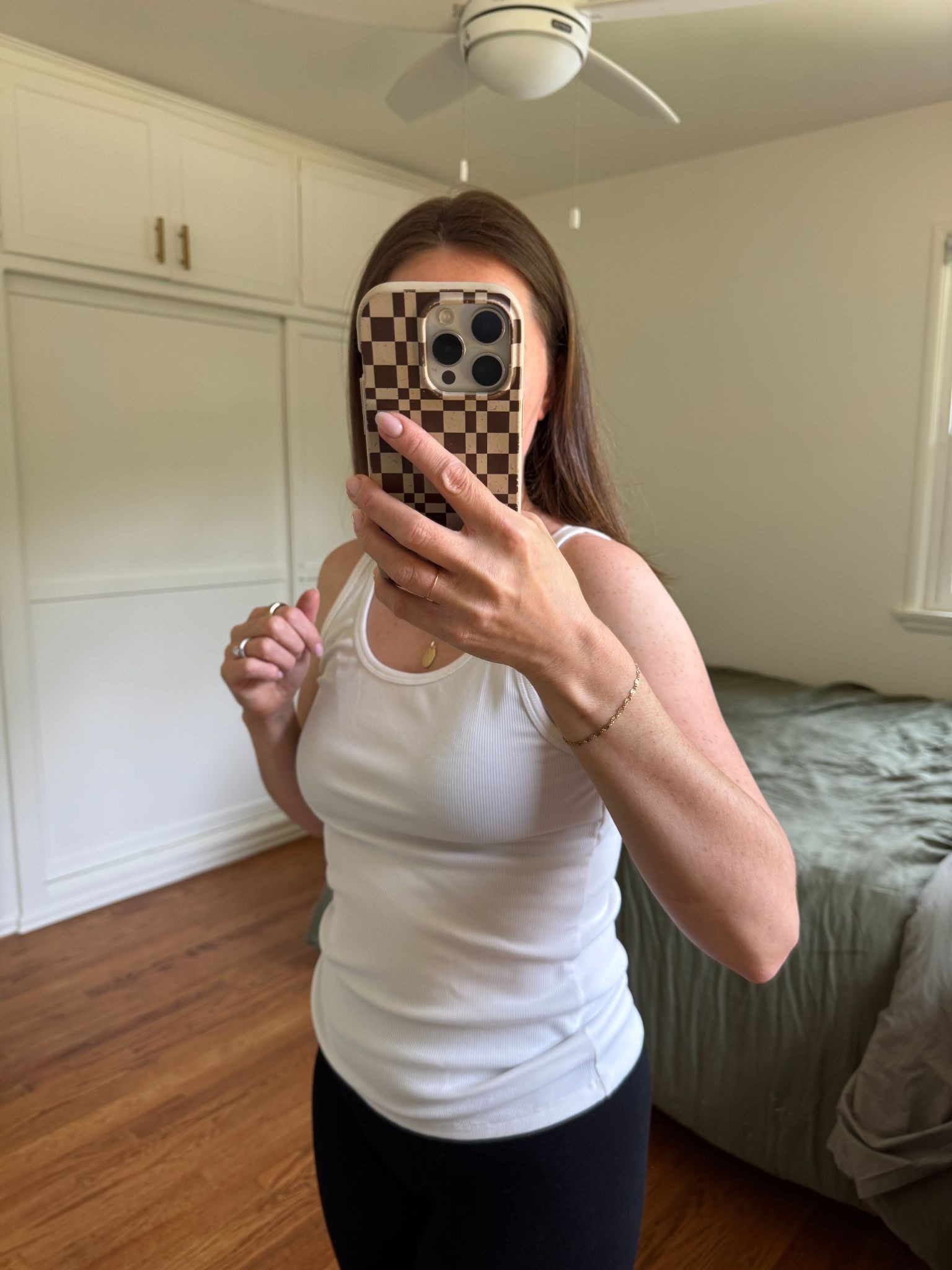 A person takes a mirror selfie in a bedroom, wearing a white tank top and black pants, holding a phone with a checkered case.