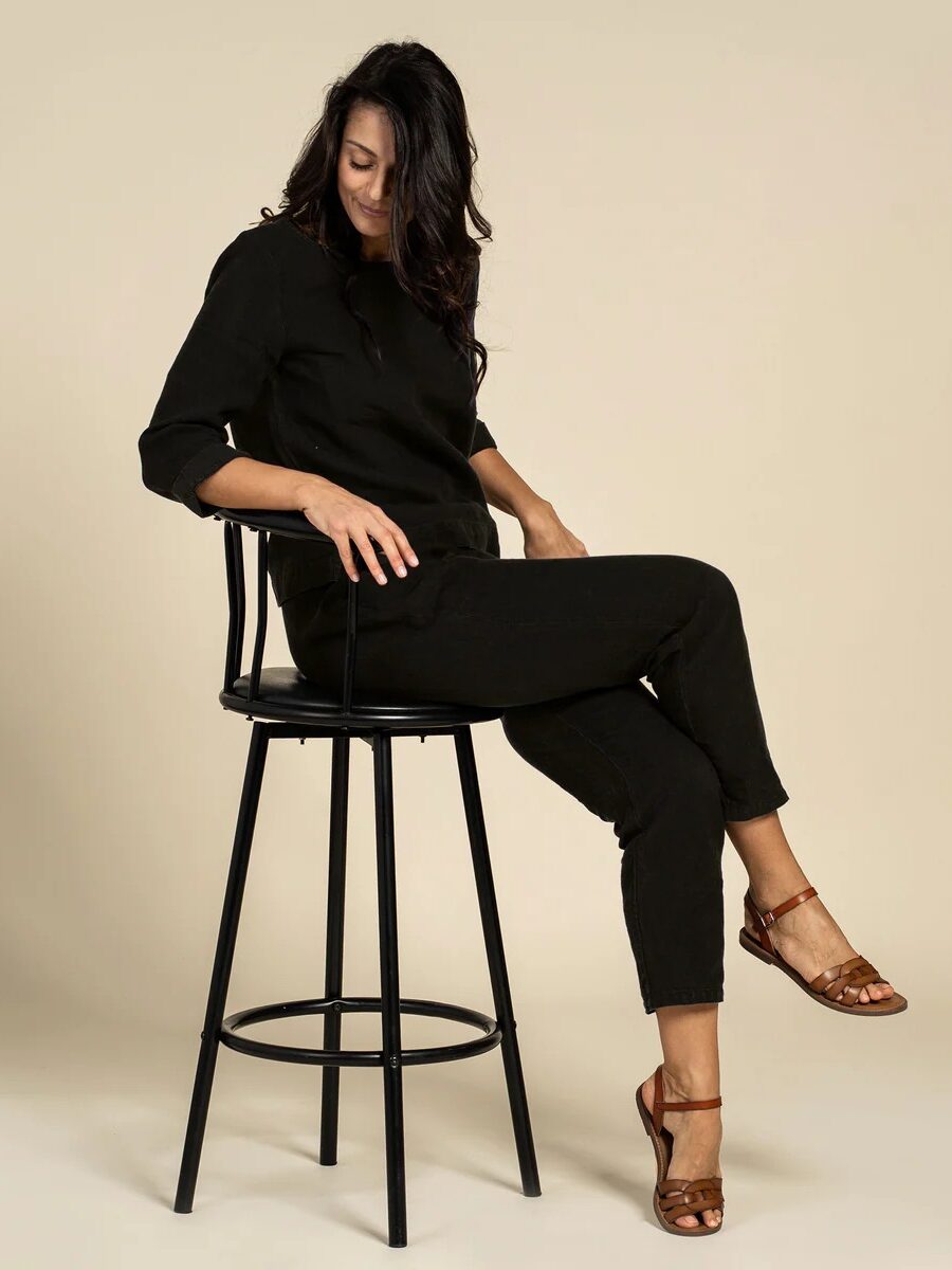 A woman with long dark hair sits sideways on a tall black stool, wearing a black outfit and brown sandals, looking down with a neutral expression against a beige background.