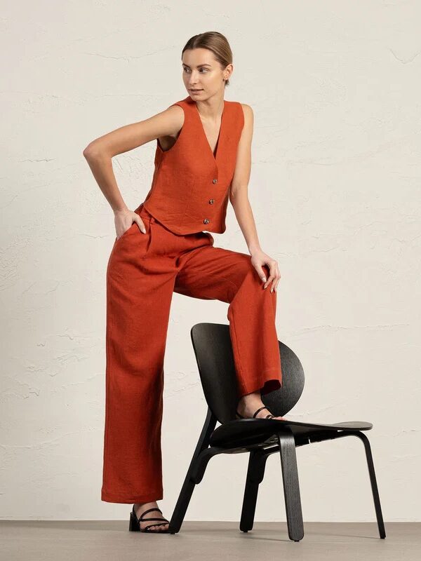 A woman wearing a sleeveless rust-colored vest and matching wide-leg pants poses with one foot on a black chair against a plain light background.