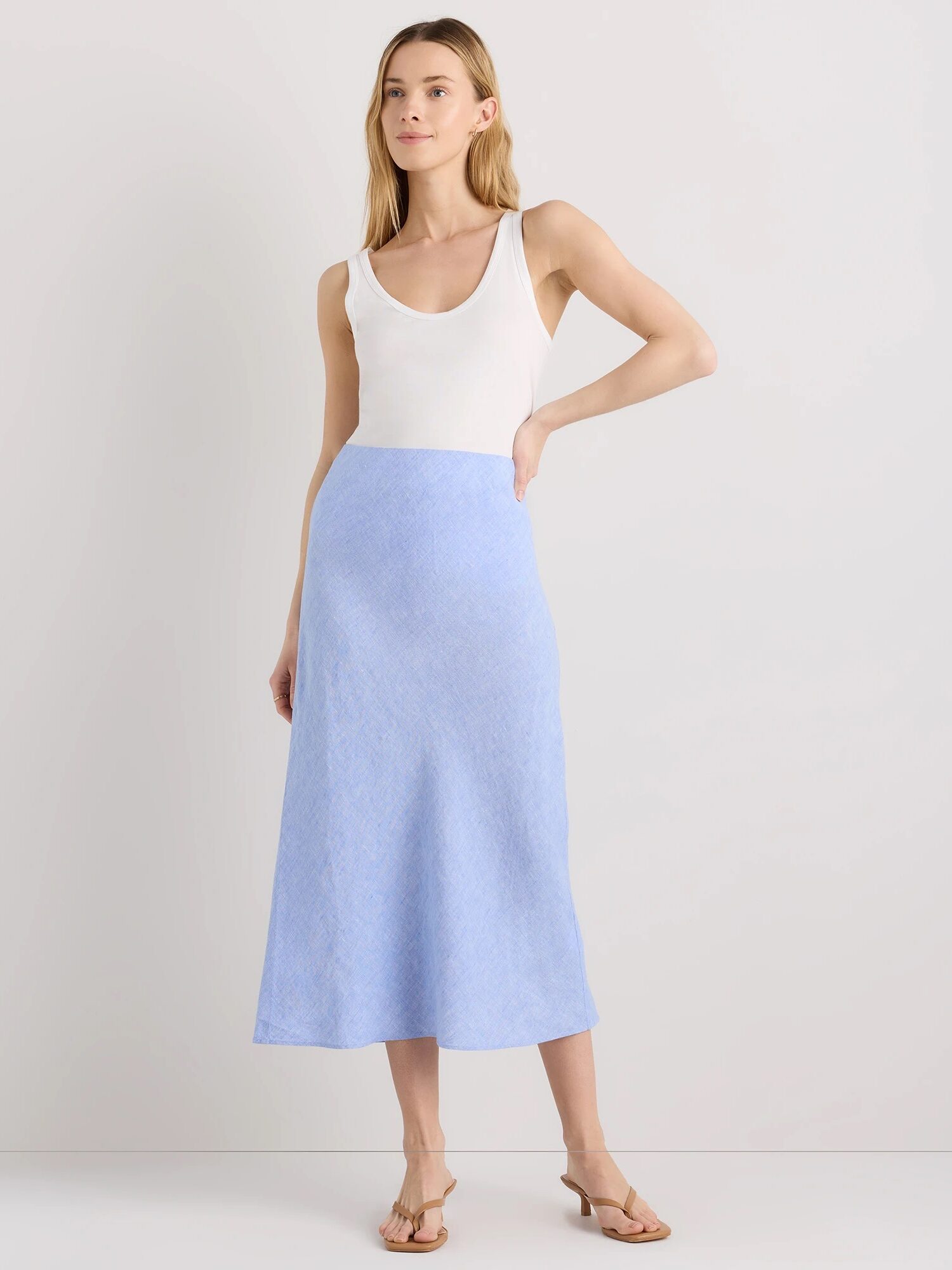 A woman stands against a plain background wearing a white sleeveless top, a light blue ankle-length skirt, and beige heeled sandals.