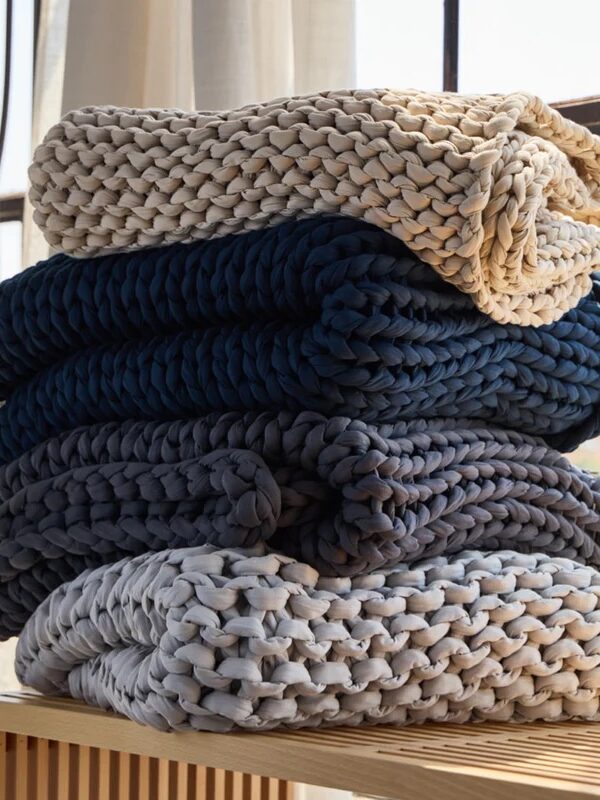 A stack of four chunky knit blankets in beige, navy blue, gray, and light gray, folded and placed on a wooden surface near a window.