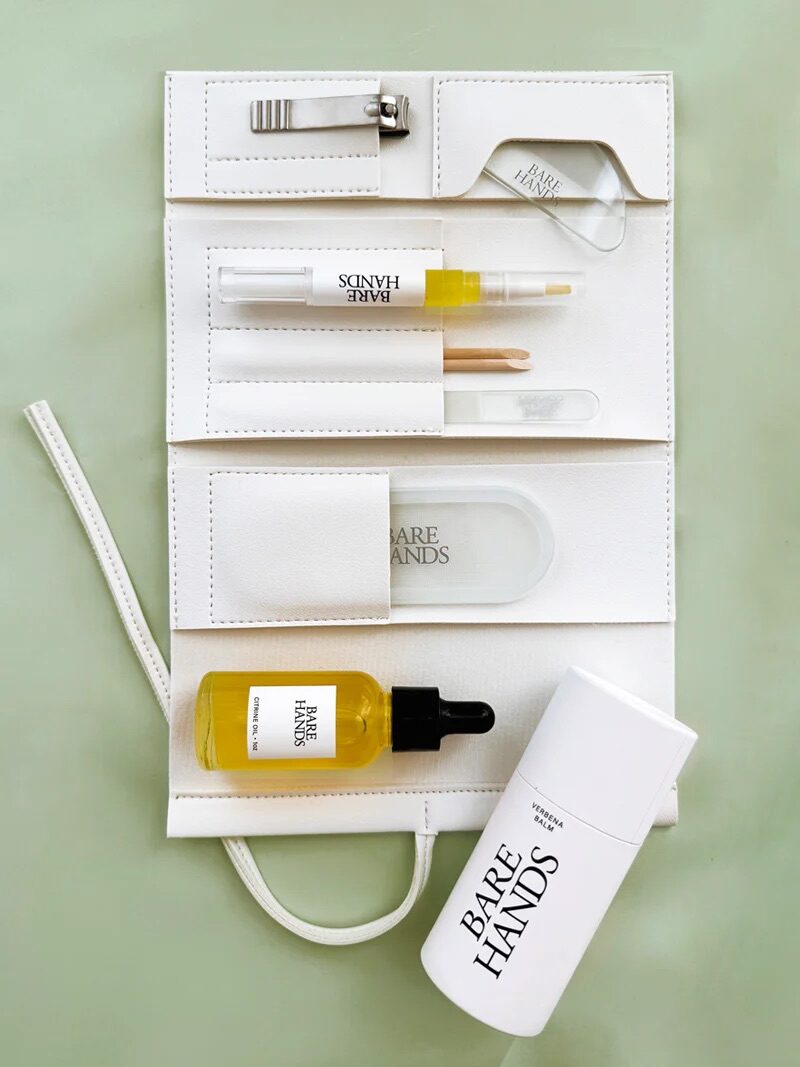A white manicure kit is laid open on a light green surface, displaying nail clippers, a glass nail file, oil, a cuticle stick, and labeled Bare Hands products.