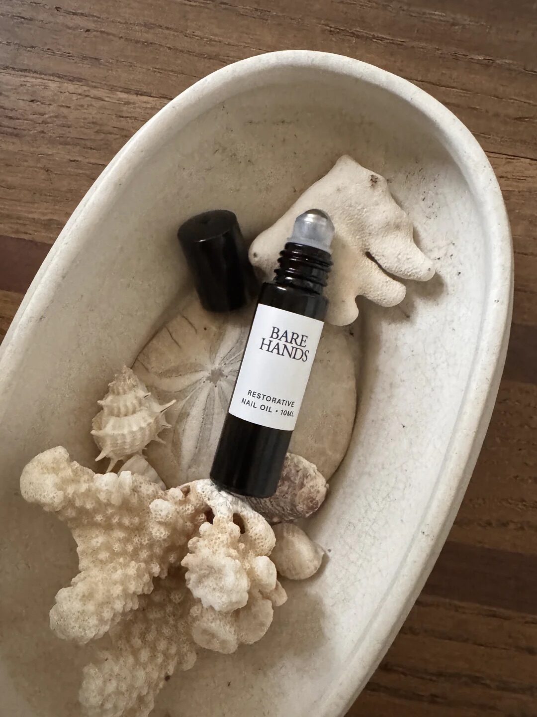 A bottle of Bare Hands restorative nail oil with a roller top is placed in a white dish with coral pieces and a sand dollar on a wooden surface.