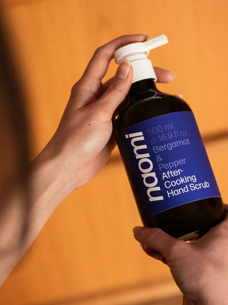 A person holds a bottle labeled "naomi Bergamot & Pepper After-Cooking Hand Scrub, 500 ml (16.9 fl oz)" with a pump dispenser.