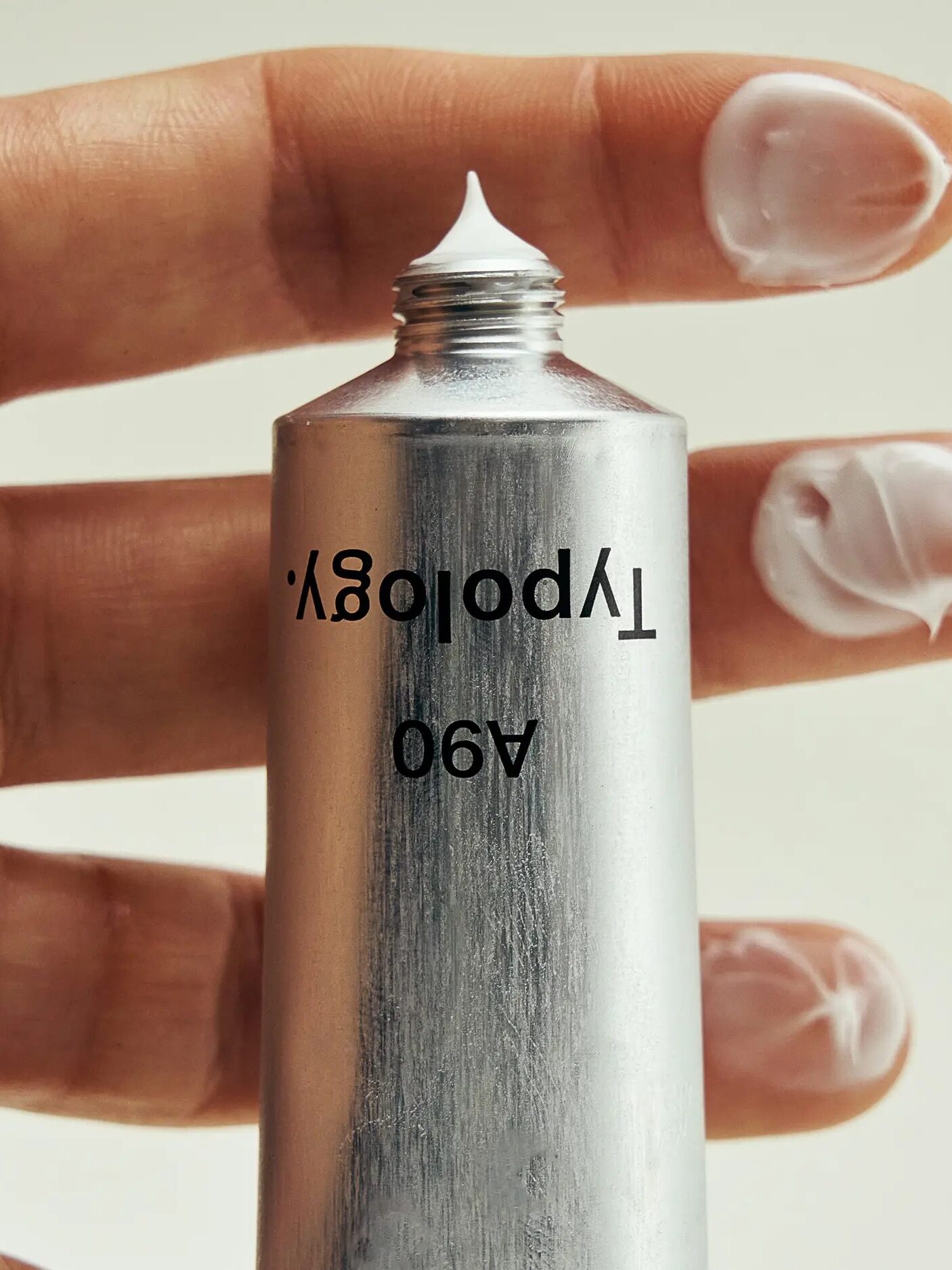 A hand with three fingers holding a metal tube with white cream; some cream is on each fingertip. The text on the tube is partially visible and upside down.