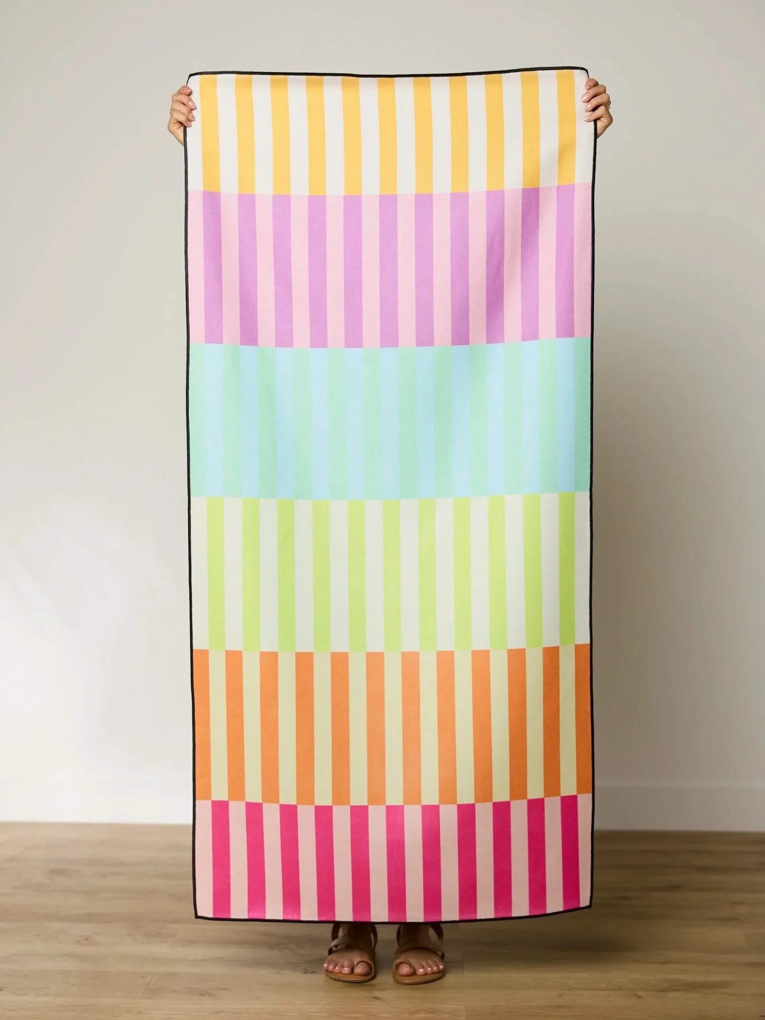 Person holding a rectangular towel with vertical stripes in pastel yellow, orange, pink, blue, green, and red, standing indoors against a plain wall.