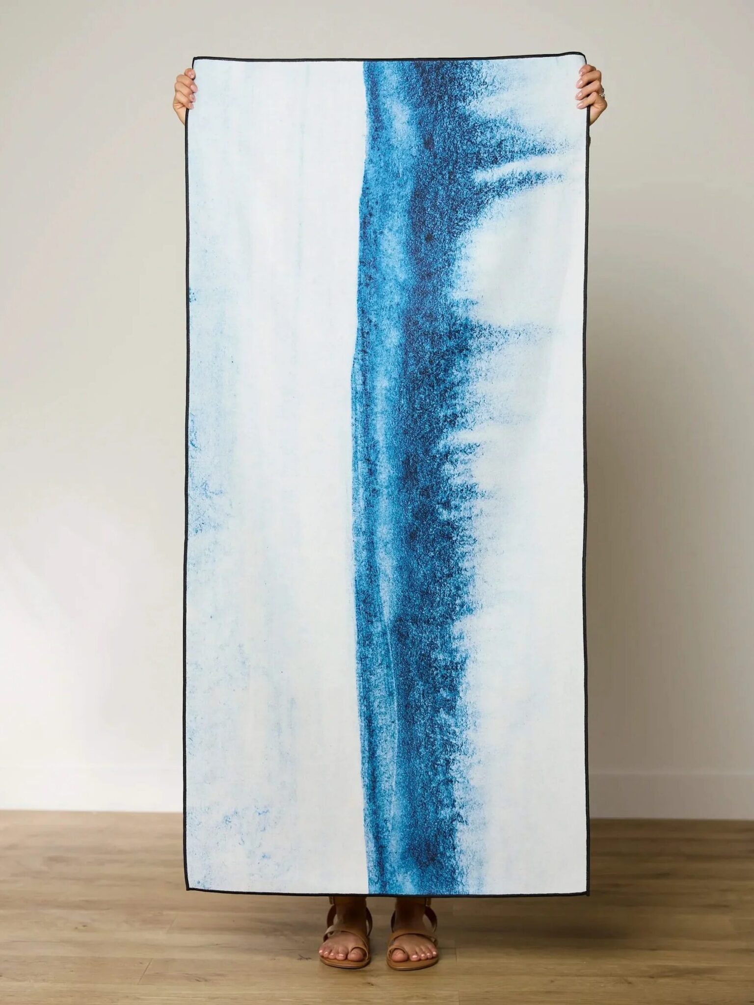 Person holding a rectangular towel vertically with a blue abstract stripe running down the center, standing against a plain wall and wooden floor.