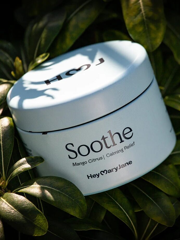 A round, light blue container labeled "Soothe" rests on green leaves, with sunlight casting shadows on the lid. The product is from HeyMaryJane and described as calming relief.