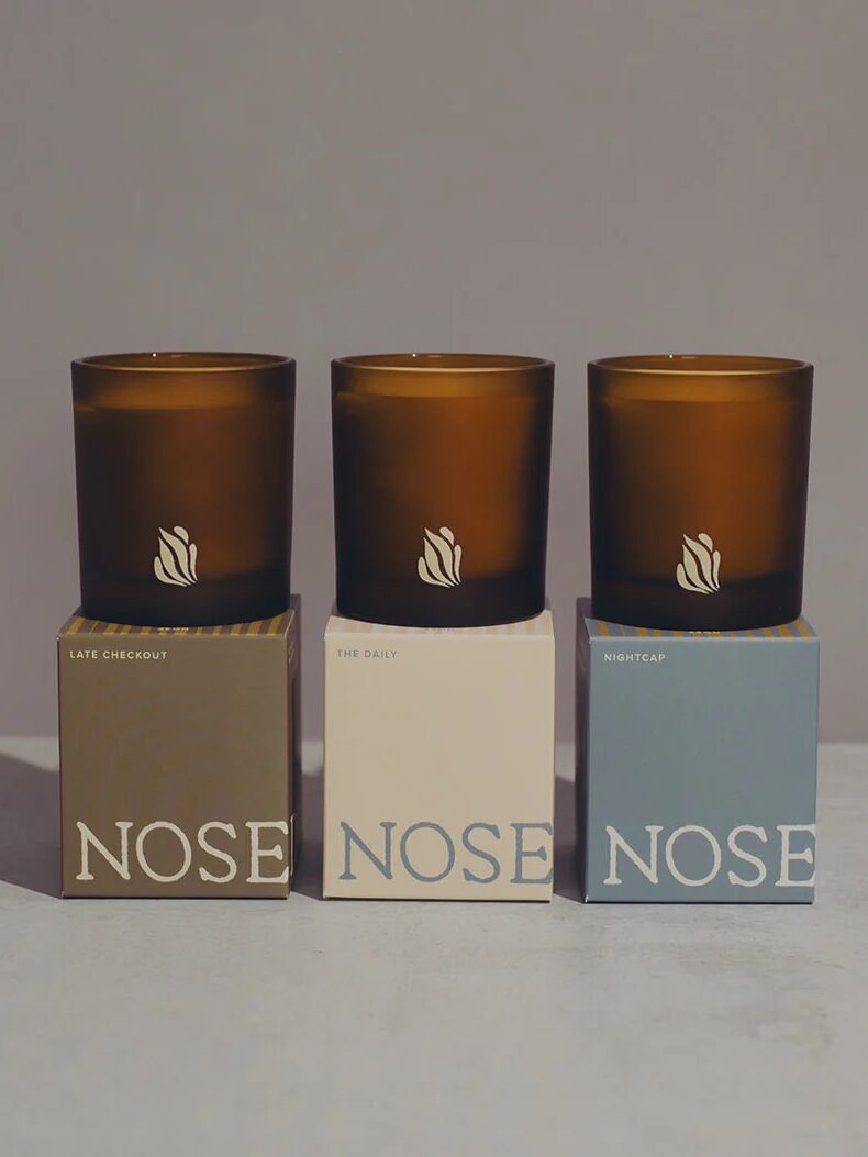 Three brown glass candles labeled "Late Checkout," "The Daily," and "Nightcap" sit atop matching boxes with the brand name "NOSE" printed on each box.