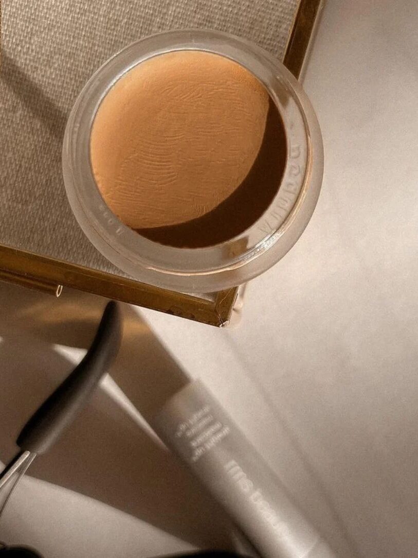 A glass container of tan cream makeup sits on a beige surface next to a partially visible tube and black-handled tool.