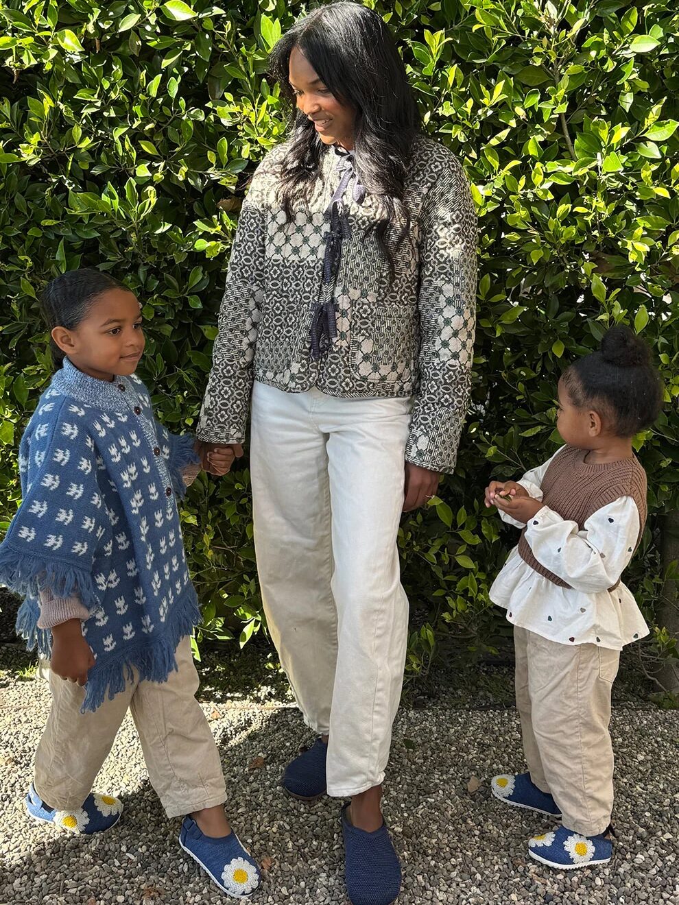 An adult and two children stand on gravel in front of green bushes, wearing casual outfits with patterned sweaters and blue slip-on shoes.