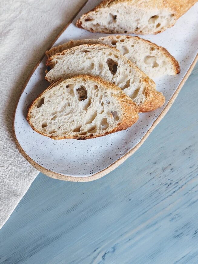 Three slices of artisanal bread are arranged on a speckled white ceramic plate, placed on a blue wooden surface next to a light-colored cloth.