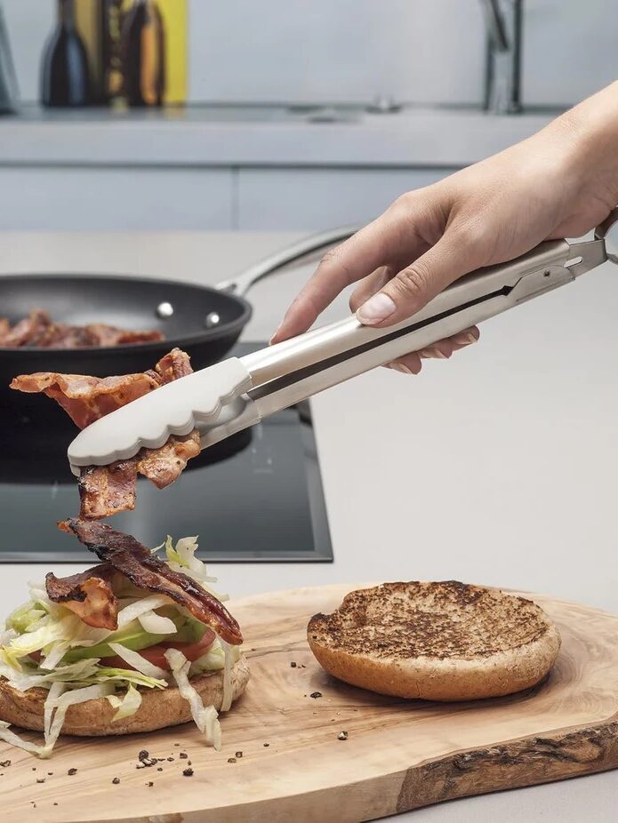 A hand uses metal tongs to place cooked bacon onto a sandwich with lettuce and tomato on a wooden board; an empty bun and a frying pan are nearby.