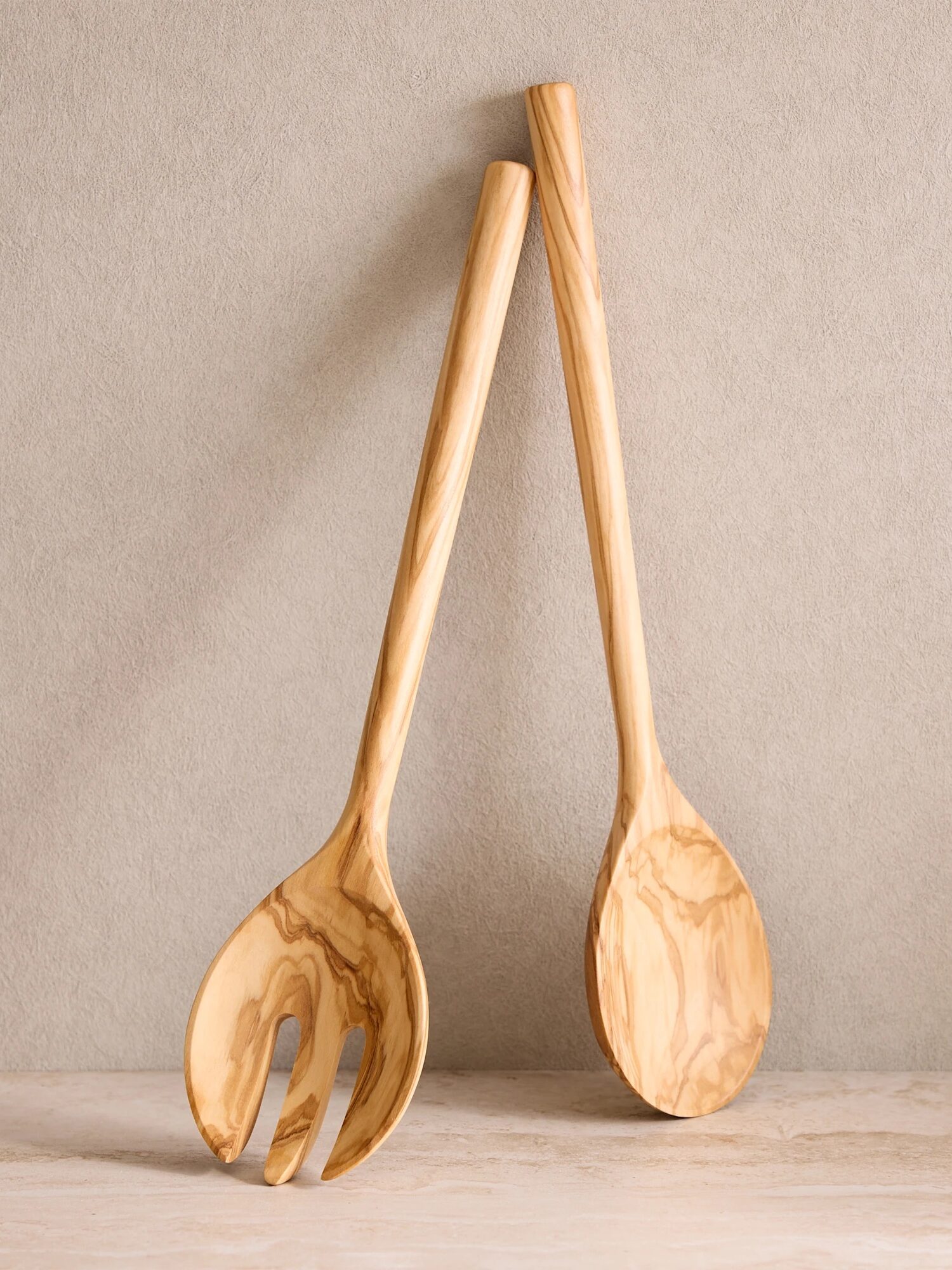 Two wooden utensils, a salad fork and a spoon, are standing upright and crossed against a neutral textured background.