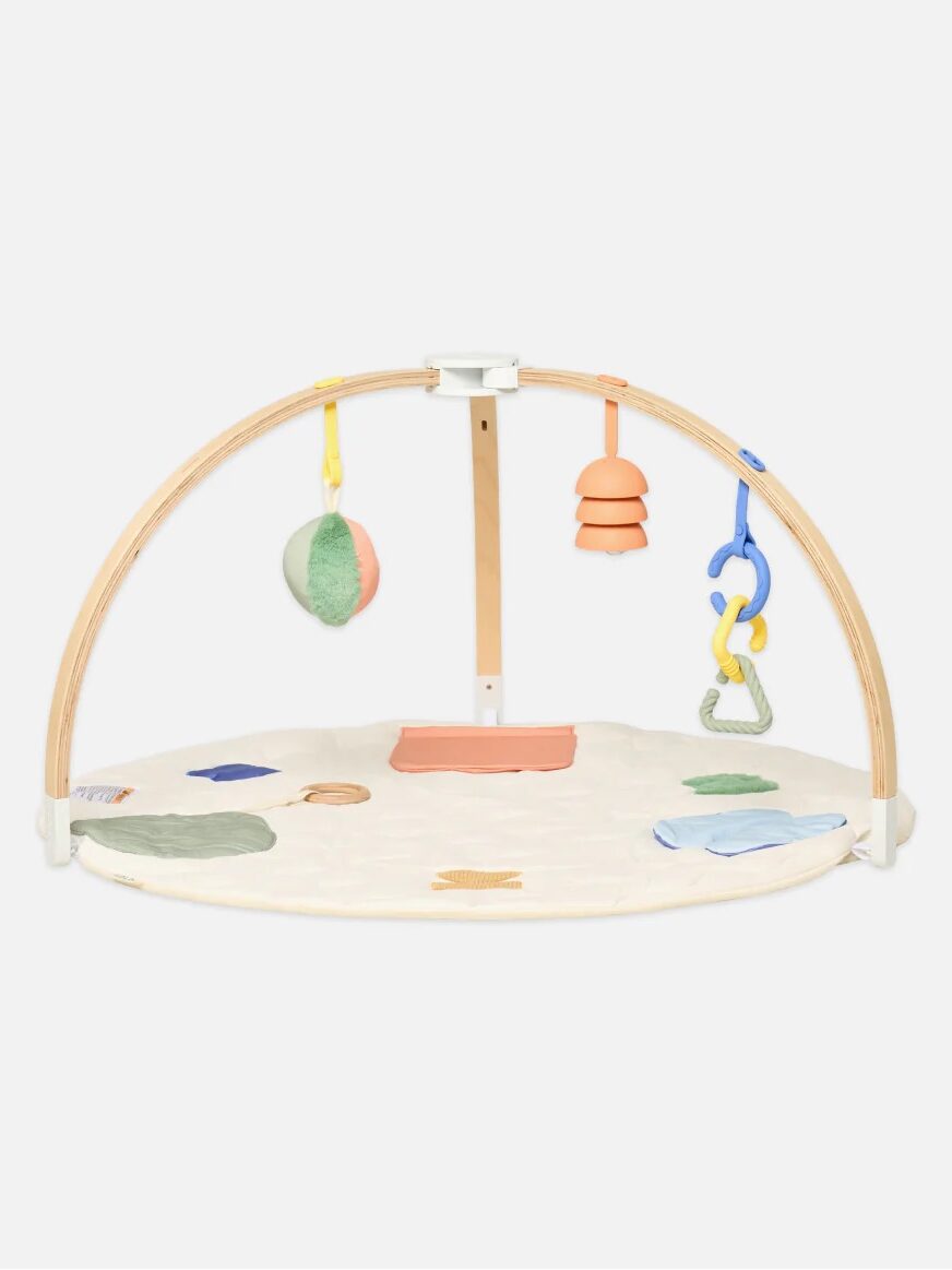 A baby play gym with a round padded mat, two wooden arches, and hanging toys in pastel colors.