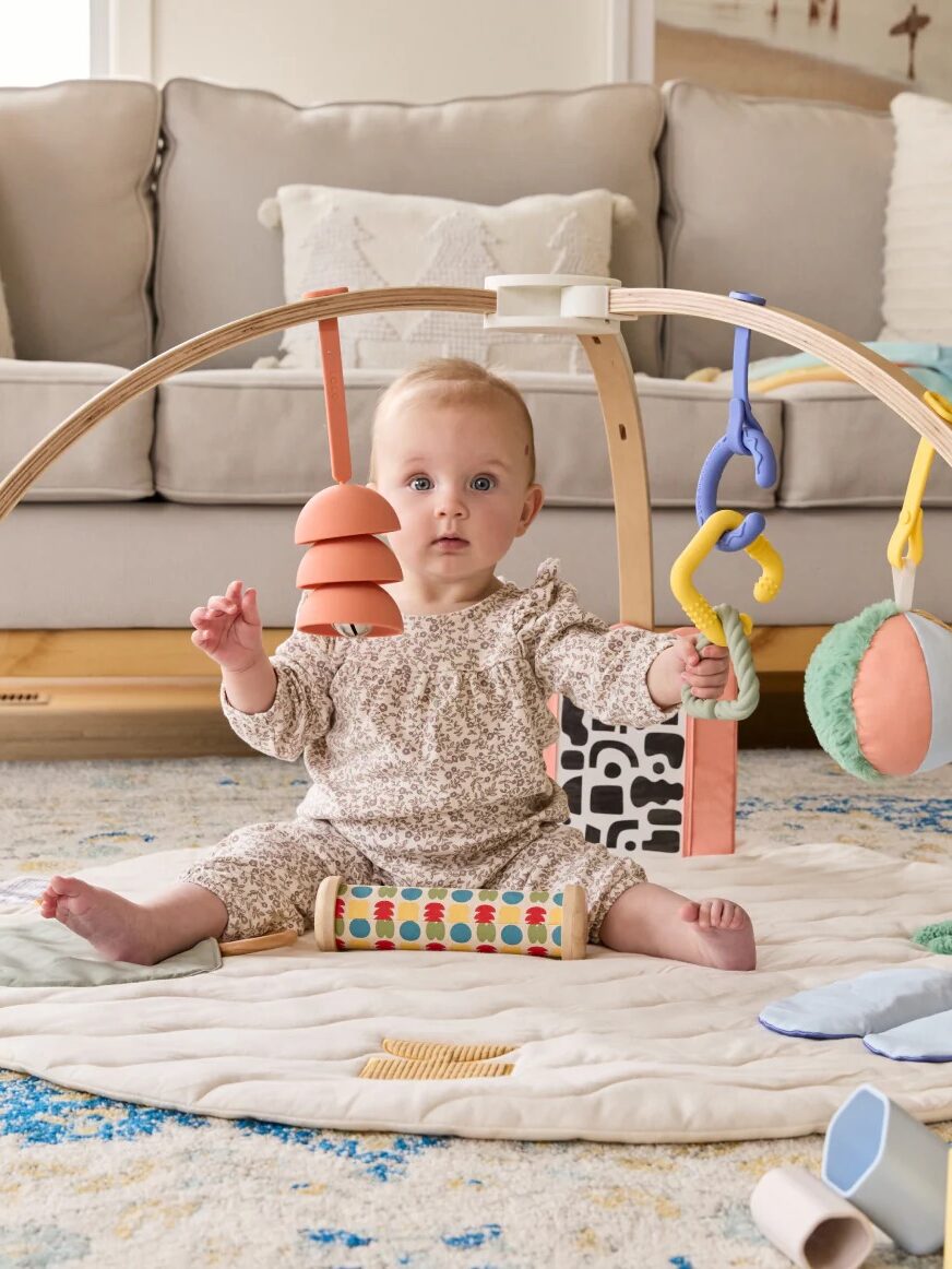 A baby sits on a play mat, surrounded by soft toys and hanging objects, in a living room with a sofa and cushions in the background.