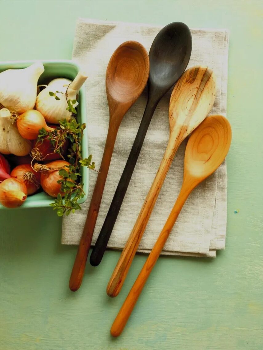 A bowl of onions and garlic sits next to four wooden spoons and a folded napkin on a light green surface.