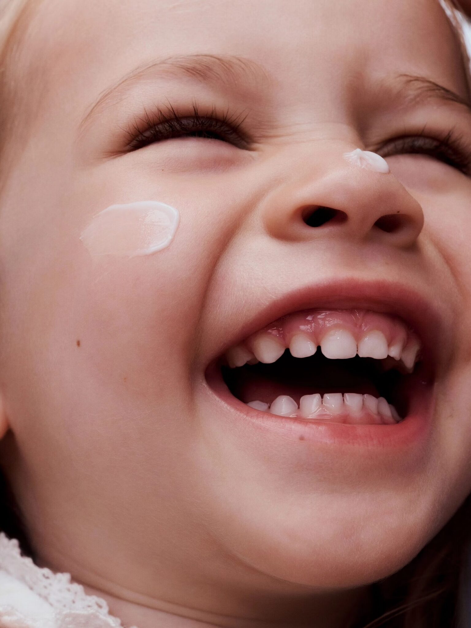 Close-up of a smiling child with an open mouth, showing teeth, and a dab of cream on their cheek.