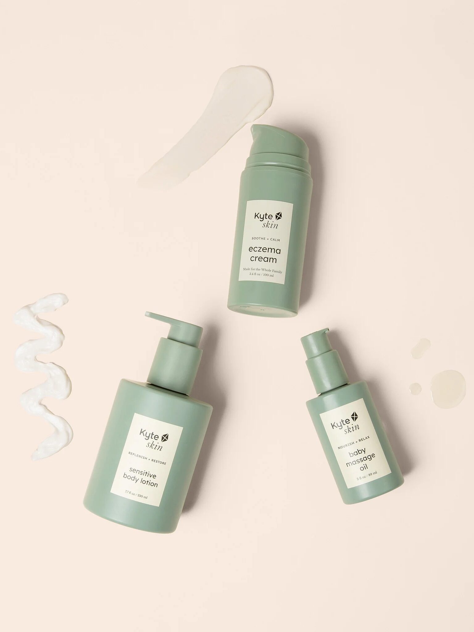 Three green skincare bottles labeled sensitive body lotion, eczema cream, and calming baby oil are arranged on a light beige surface with small product smears nearby.