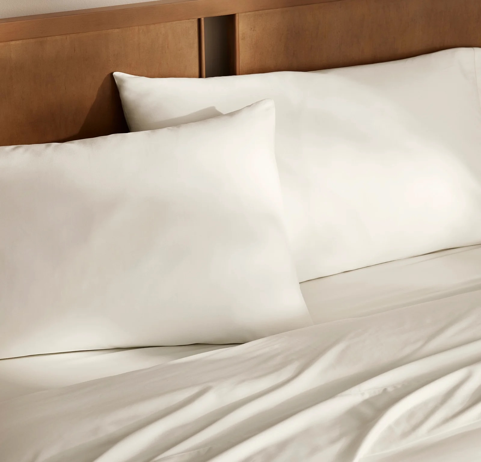 Two white pillows rest against a wooden headboard on a neatly made bed with white sheets.