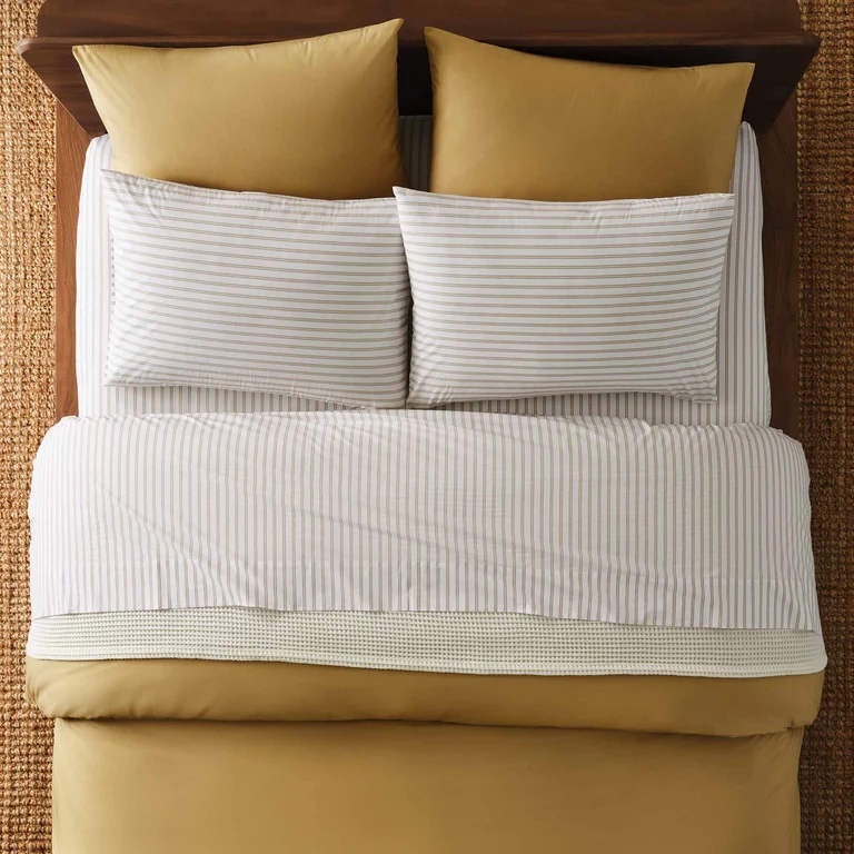 A neatly made bed with gold-colored pillows and comforter, white and gray striped sheets, and a textured woven rug underneath.