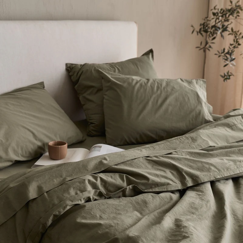 A neatly made bed with olive green sheets and pillows, an open book, and a brown cup placed on top, next to a plant in the background.