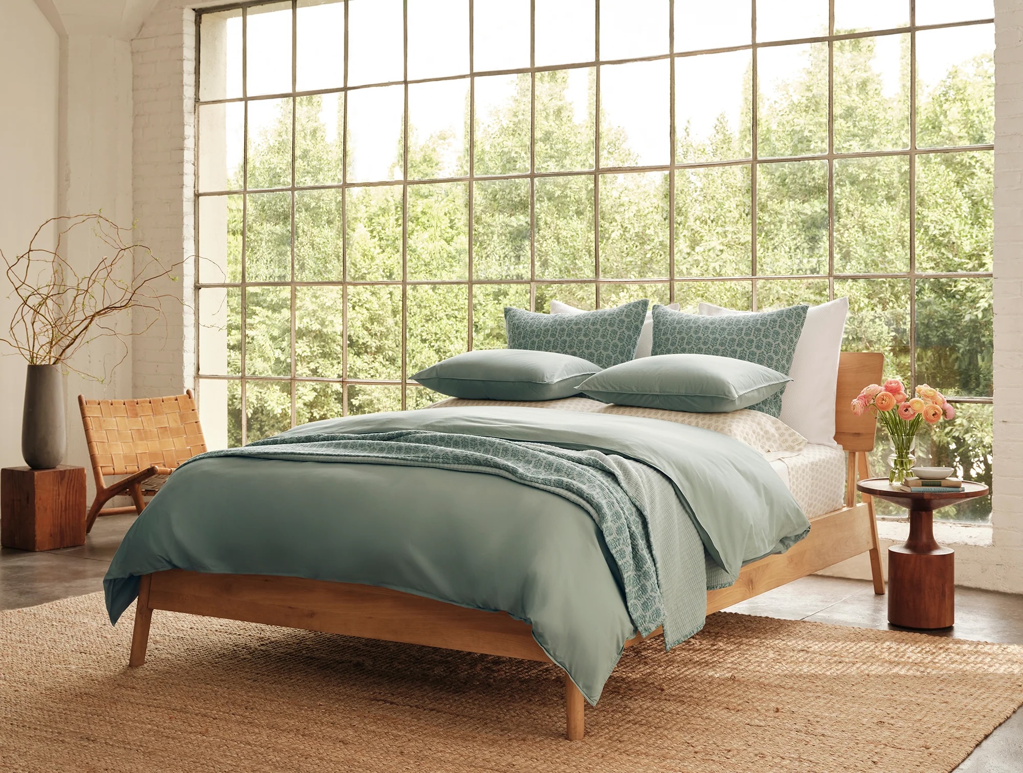 A neatly made bed with teal bedding in a sunlit room with large windows, wooden furniture, a woven rug, and a vase of flowers on a bedside table.