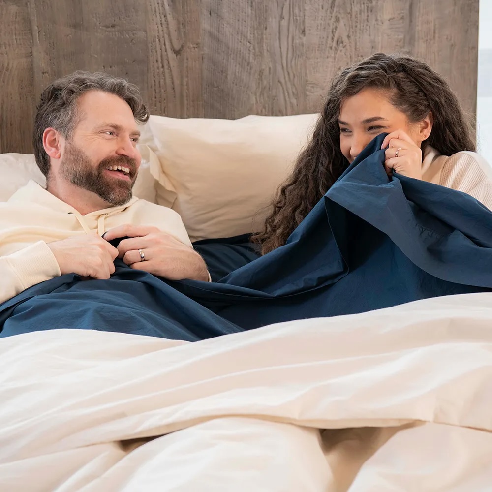 A man and woman lie in bed under navy blue and white blankets, smiling and looking at each other.