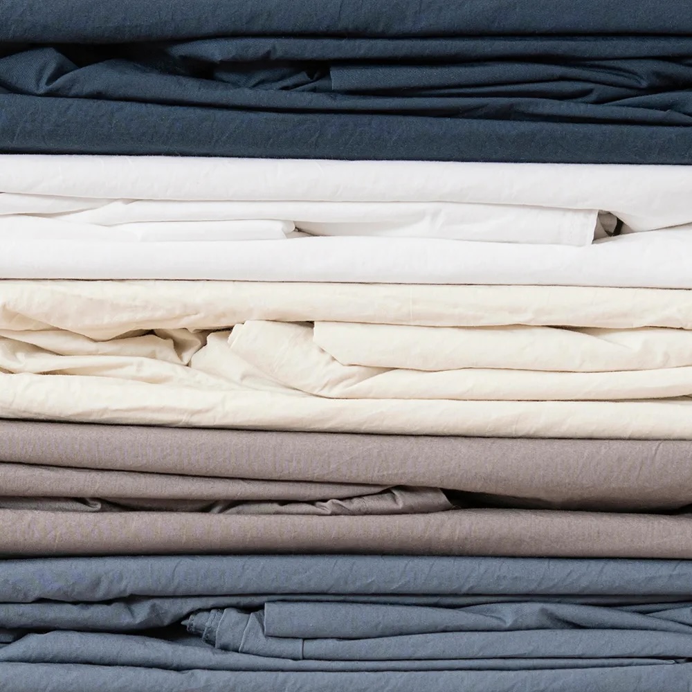 Stack of folded bed sheets in various colors, including navy blue, white, cream, grey, and light blue.