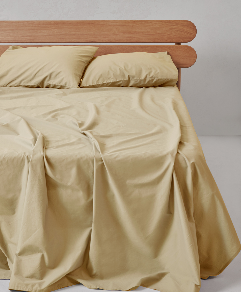 A neatly made bed with beige sheets and two pillows, featuring a wooden headboard against a plain light-colored wall.