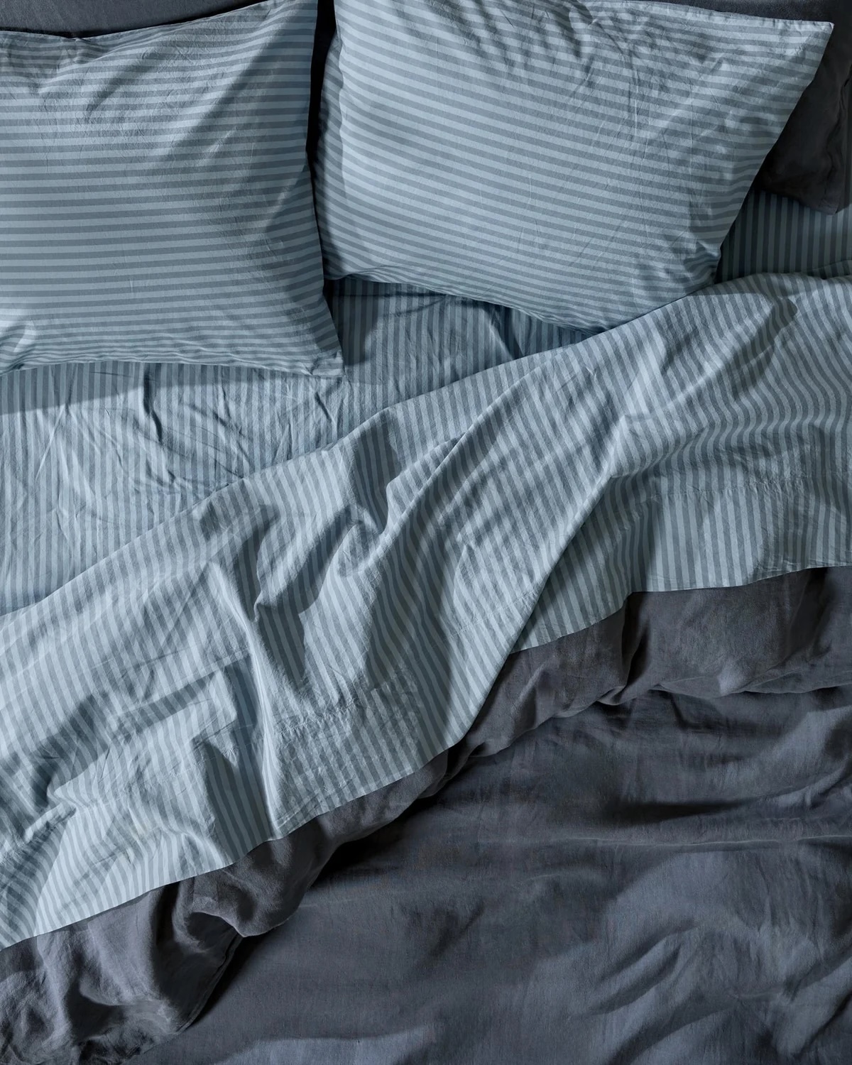 A bed with light blue and gray striped sheets and matching pillowcases appears unmade, with the bedding rumpled and draped unevenly.