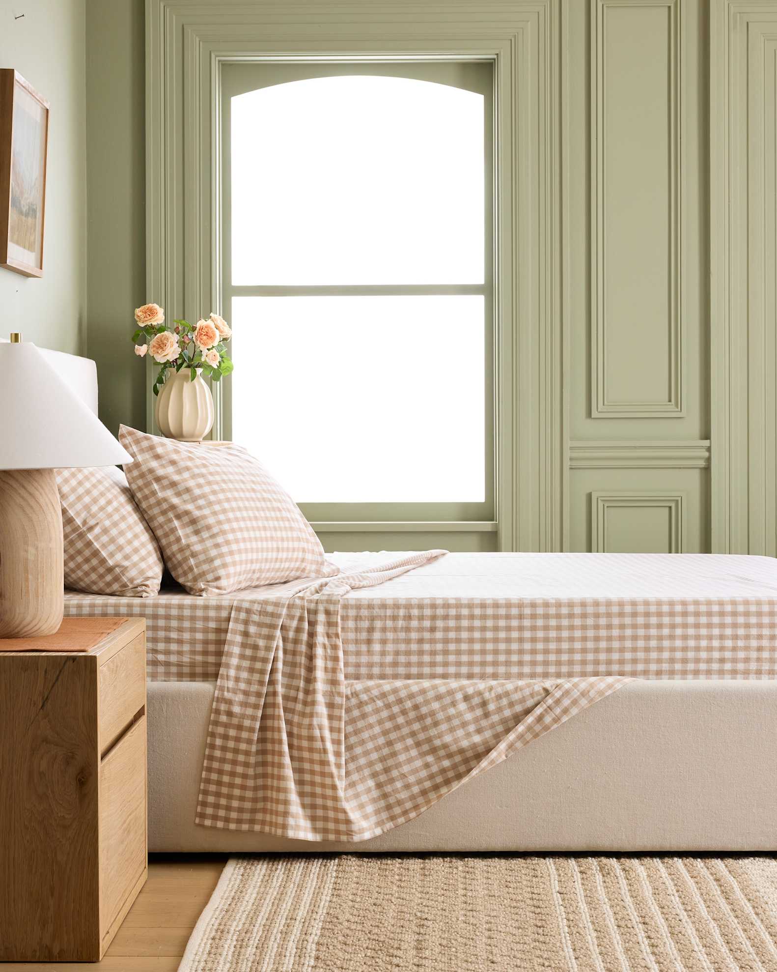 A neatly made bed with beige gingham bedding sits beside a wooden nightstand with a lamp and a vase of flowers; large window and pale green wall in the background.