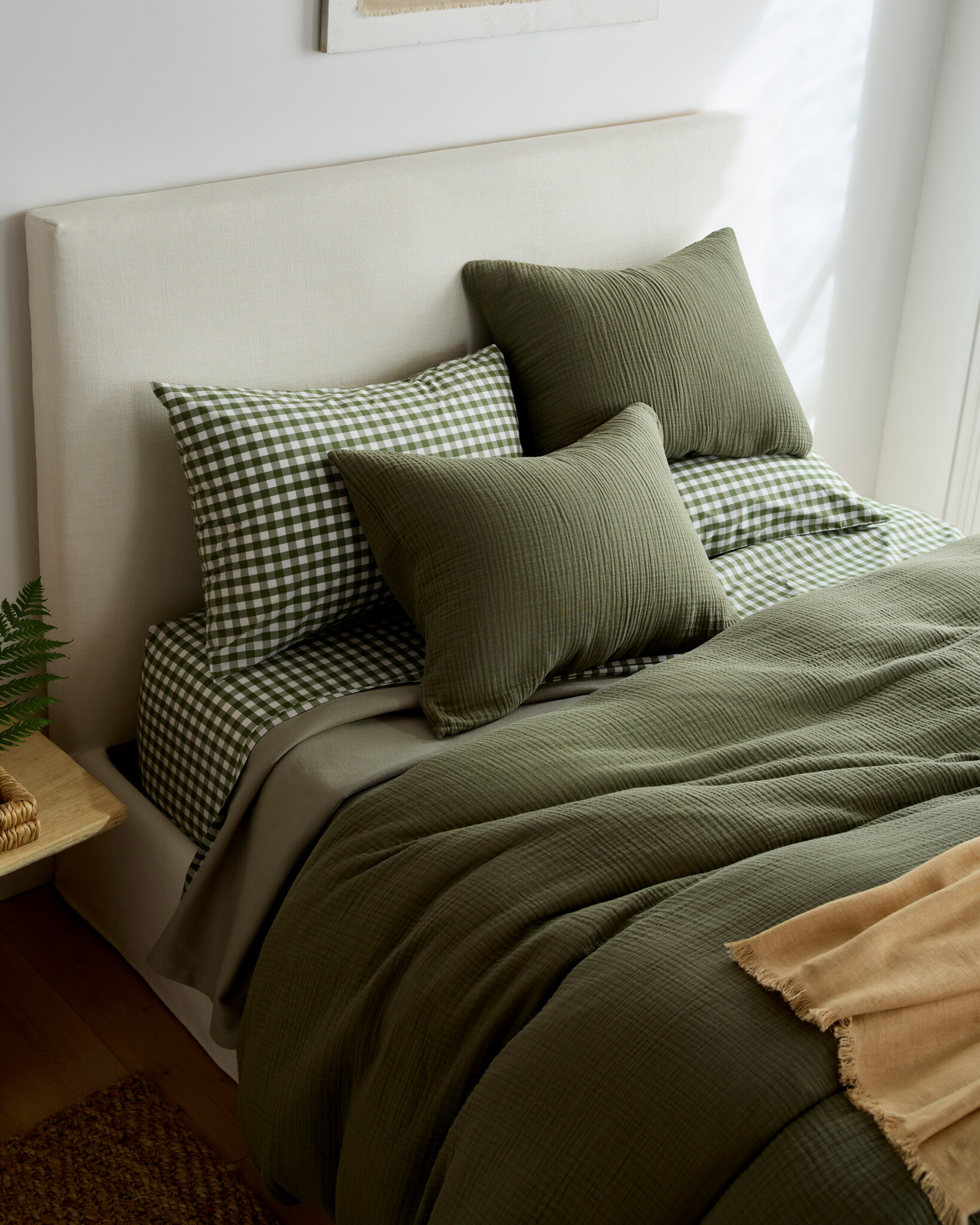 A neatly made bed with a white headboard, green checkered pillows, solid green pillows and bedding, and a beige throw blanket at the foot of the bed.