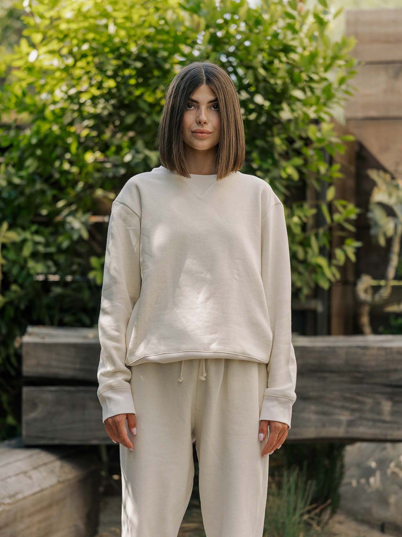 A person with straight, shoulder-length hair stands outdoors wearing a loose-fitting, light beige sweatshirt and matching sweatpants. Green foliage and wooden beams are in the background.