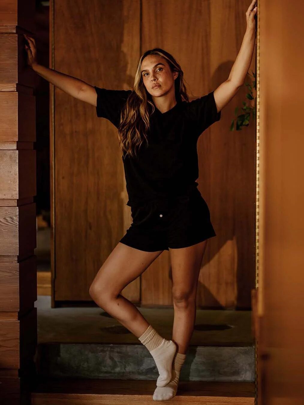 A person stands in a doorway with one arm raised, wearing a black t-shirt, black shorts, and mismatched socks in a warmly lit wooden interior.