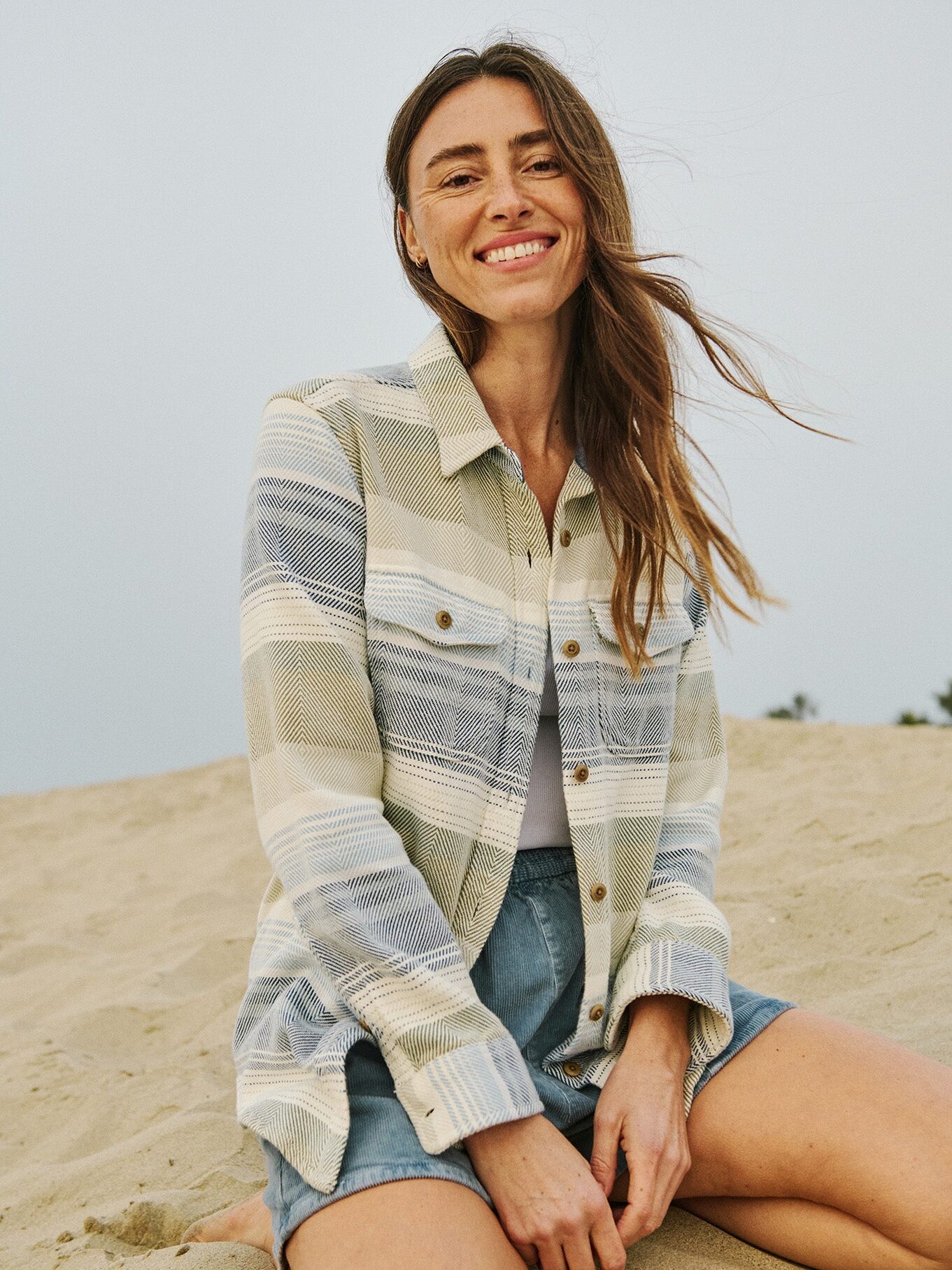 A woman sits on sand, smiling at the camera. She wears a light plaid jacket over a white top and denim shorts. Trees and a pale sky are visible in the background.