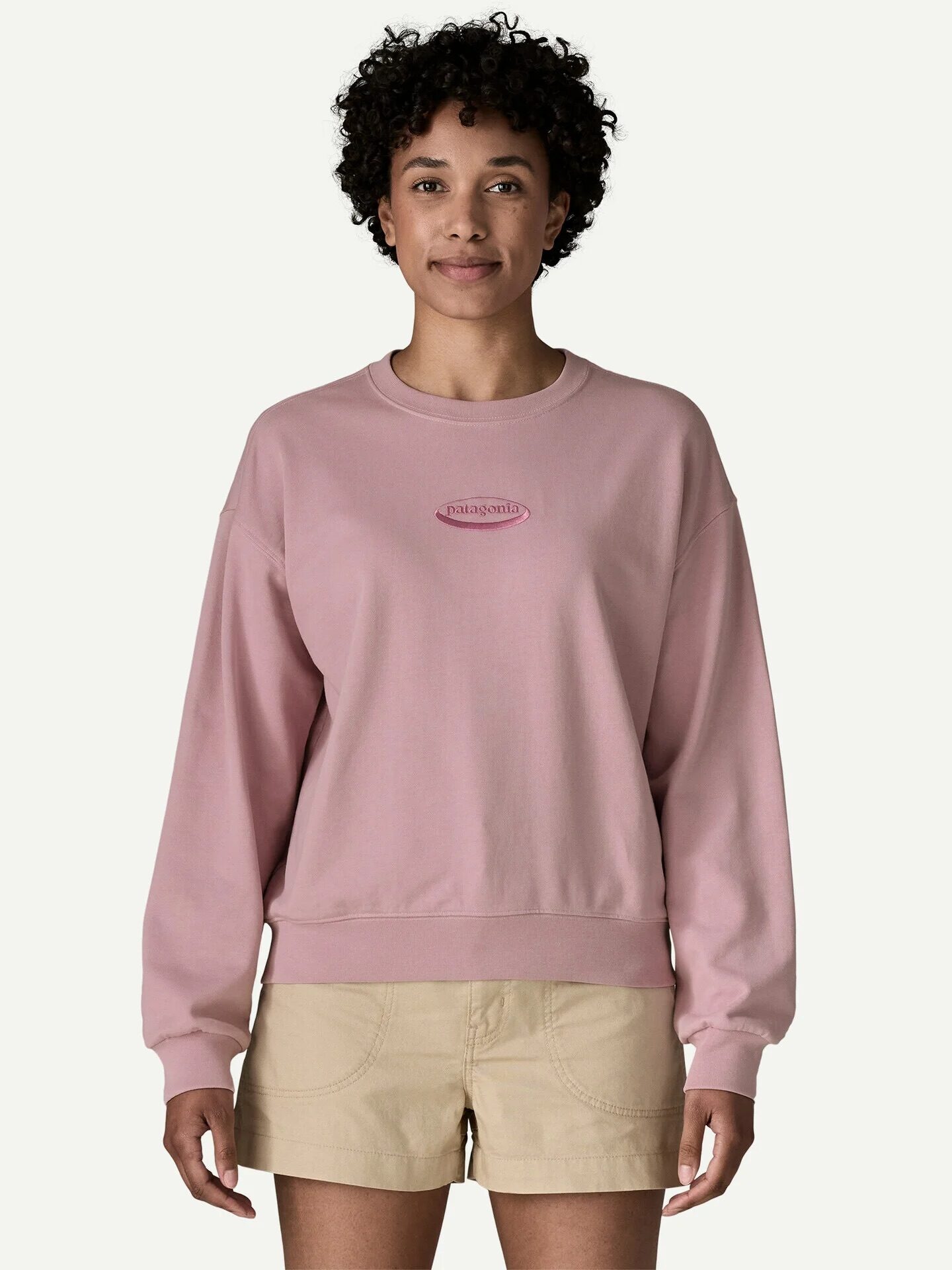 A person wearing a light pink sweatshirt with a small logo and beige shorts stands facing forward against a plain light background.