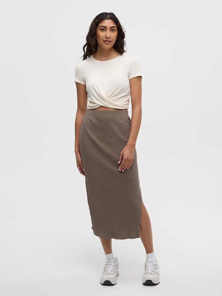 A woman stands against a plain background wearing a white knotted short-sleeve top, a ribbed taupe midi skirt with a side slit, and white sneakers.