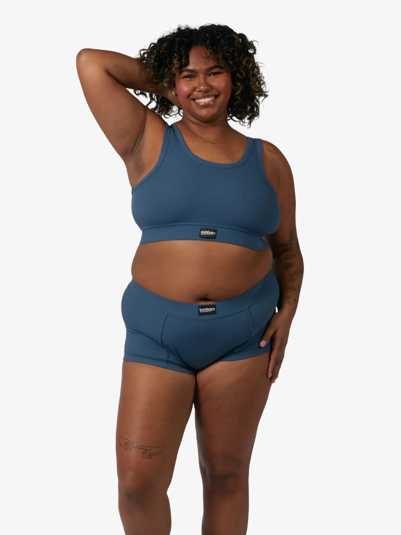 A person with curly hair poses smiling, wearing a matching blue sports bra and high-waisted underwear set against a plain white background.