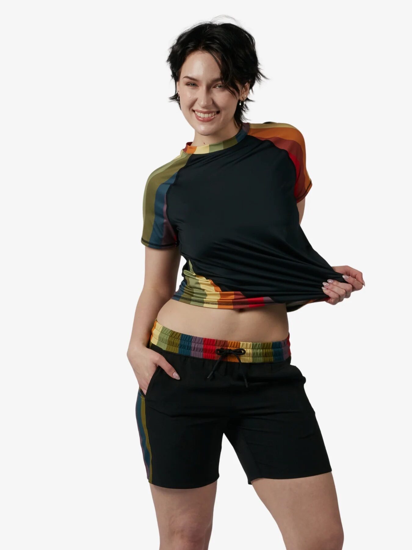 Person wearing a black rashguard and matching shorts with rainbow-colored accents, standing against a white background and smiling while holding the hem of the shirt.