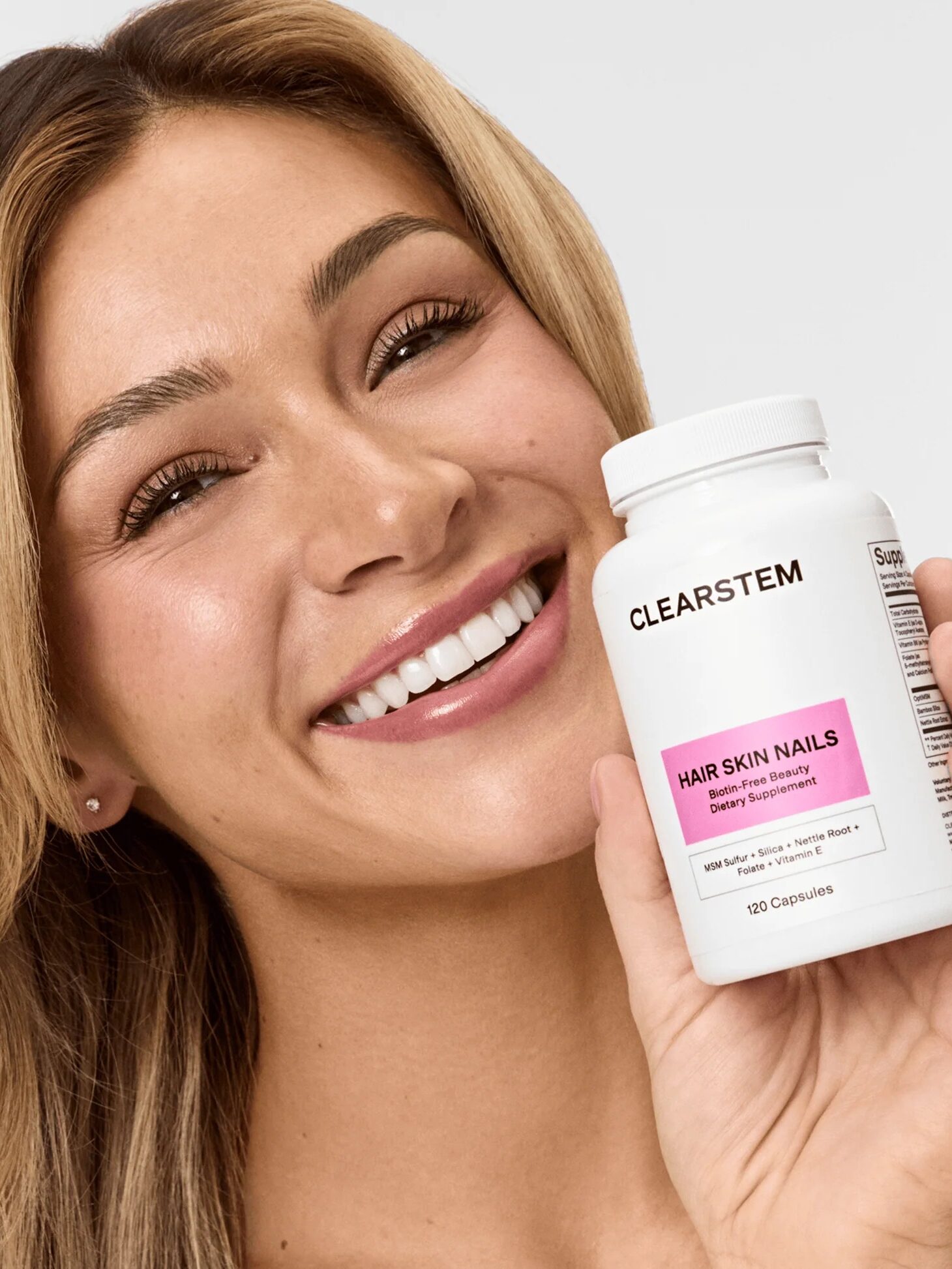 Smiling person holding a white bottle of CLEARSTEM Hair Skin Nails dietary supplement with a pink label, against a plain background.