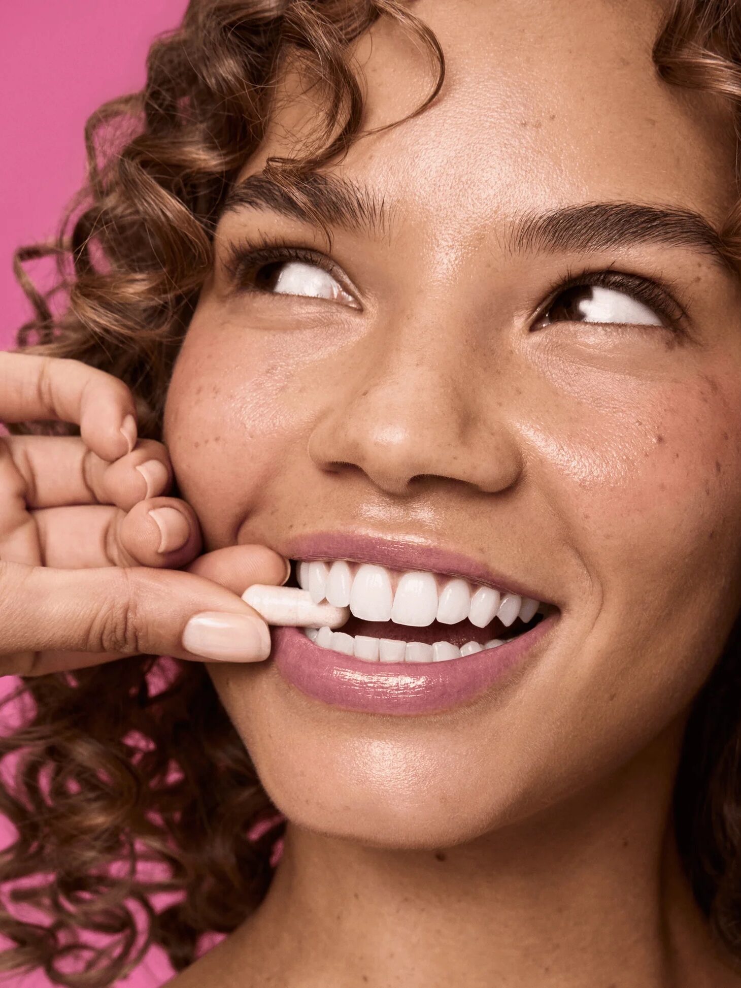 A person with curly hair smiles while another hand places a white dental strip on their upper teeth against a pink background.