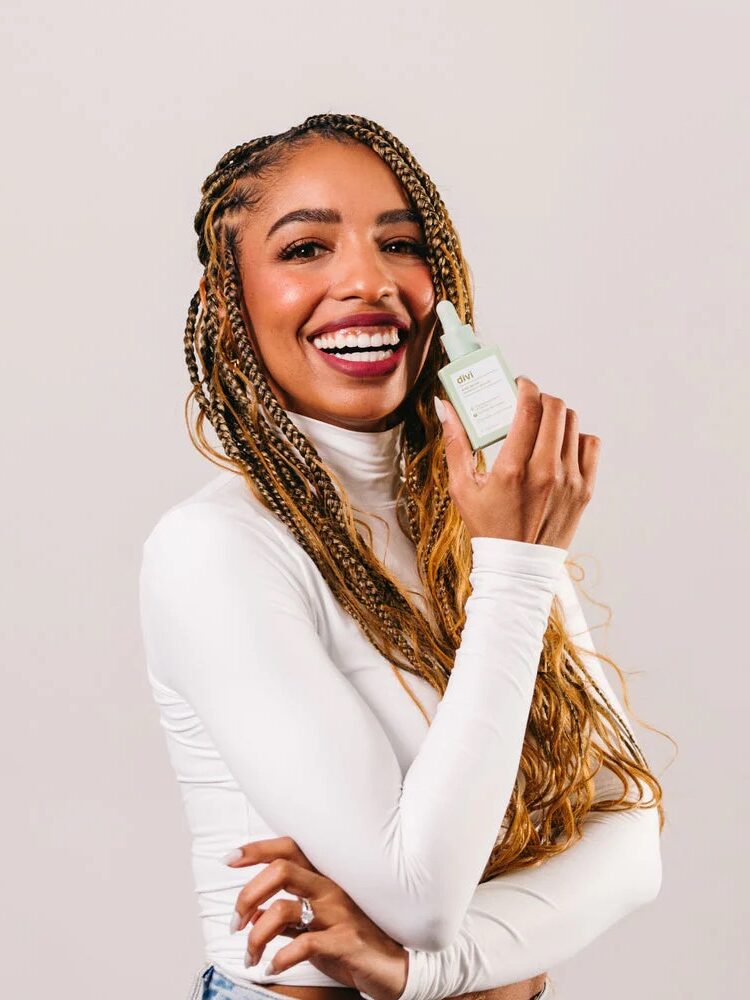 A smiling woman with long braided hair wears a white turtleneck and holds a small skincare product bottle near her face against a plain background.