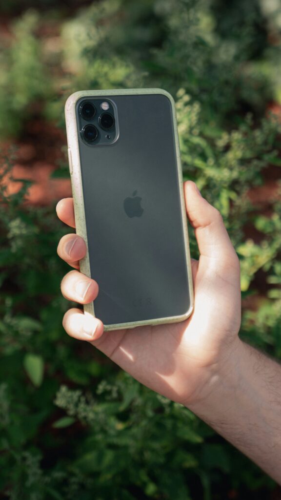A hand holding a black iPhone with a clear case outdoors, with green plants blurred in the background.
