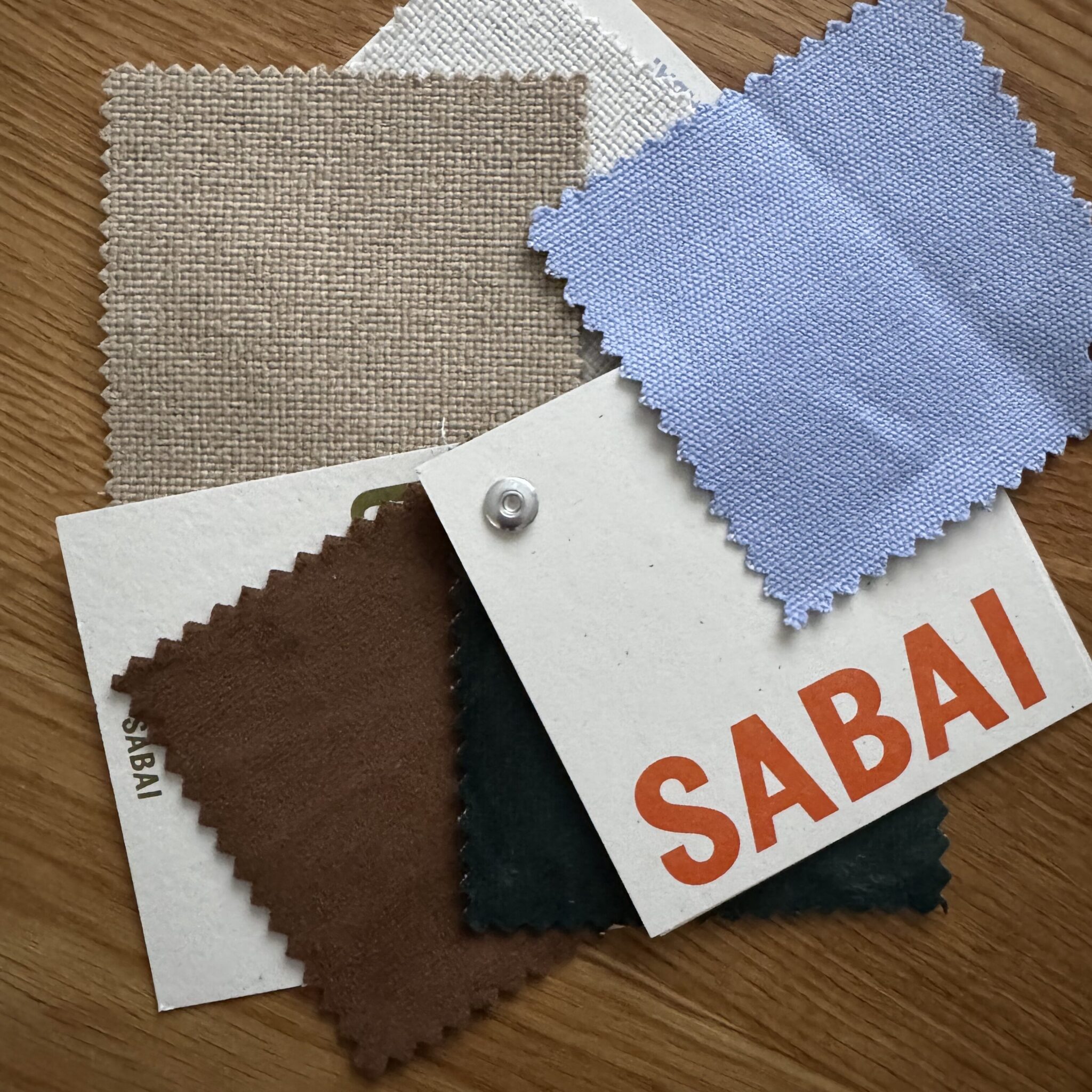 Five fabric swatches in neutral and blue tones, one labeled "SABAI," arranged on a wood surface.