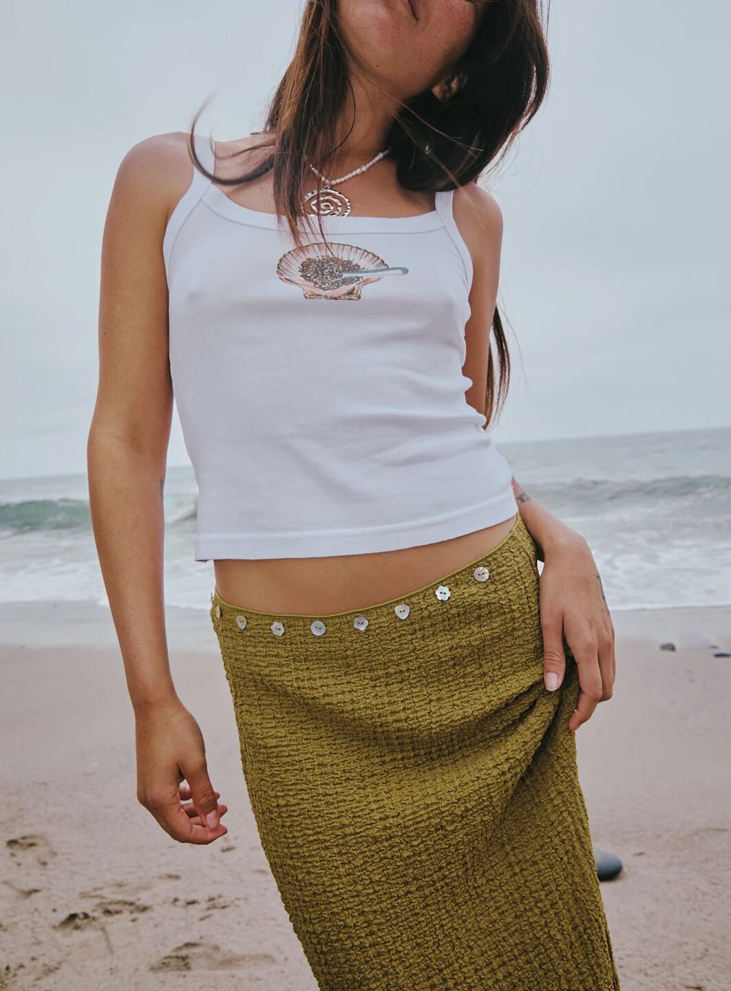 A person stands on a beach wearing a white tank top with a shell design and an olive green textured skirt with decorative buttons at the waist.