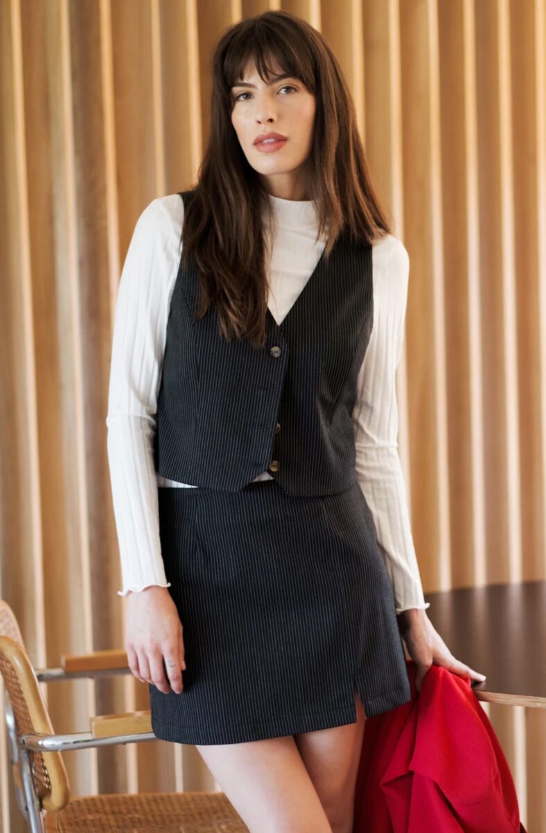 A woman stands indoors wearing a black pinstripe vest and skirt over a white turtleneck top, holding a red jacket in her hand. Wooden vertical panels form the background.