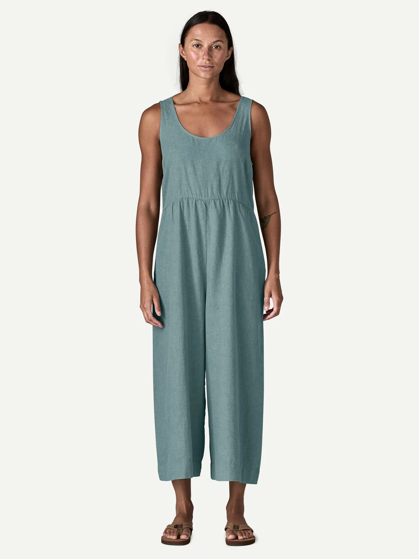 A woman stands facing forward wearing a sleeveless, teal jumpsuit with wide legs and sandals against a plain light background.